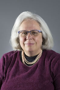 Woman wearing purple half-rim glasses. She has chin length white hair and is wearing and a purple sweater